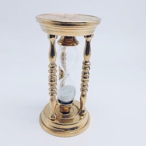 May include: A gold hourglass with a decorative base and a clear glass bulb. The hourglass is filled with white sand.