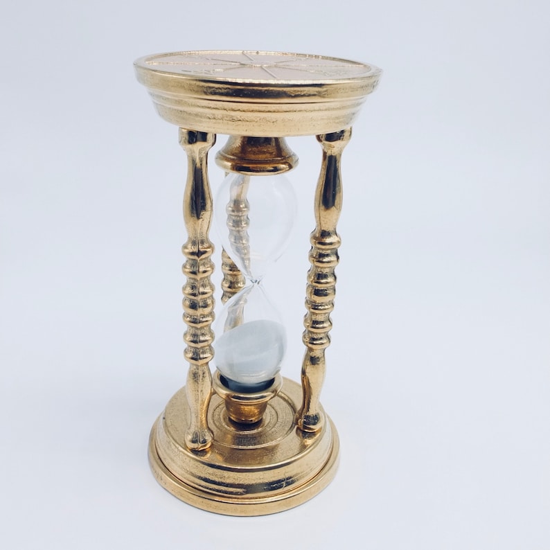 May include: A gold hourglass with a decorative base and a clear glass bulb. The hourglass is filled with white sand.