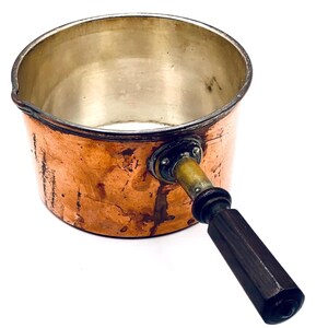 Könnte beinhalten: Ein runder Kupfertopf mit metallischer Innenauskleidung und einem dunkelbraunen, facettierten Holzgriff, der an einer kreisförmigen Metallhalterung befestigt ist. Die Außenseite des Kupfertopfes zeigt Anzeichen von natürlicher Oxidation.