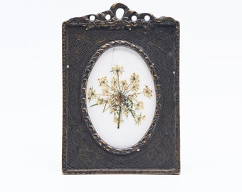 May include: A small, oval-shaped picture frame with a dark brown finish. The frame has a decorative, ornate design around the edge. A pressed flower is displayed inside the frame.