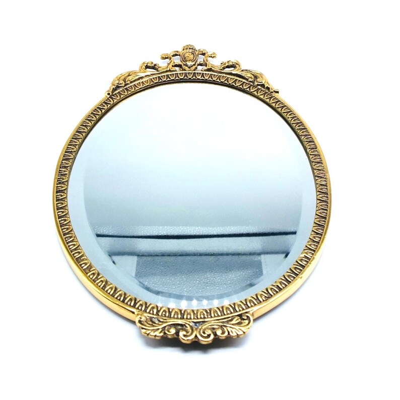 May include: A gold-toned oval hand mirror with a decorative ornate frame. The mirror has a clear glass surface.