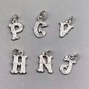 May include: Six silver alphabet charms in a vintage style, featuring the letters P, G, V, H, N, and J. Each charm has a small loop for attaching to a chain or bracelet. The charms are displayed on a grey surface.