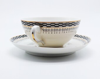 May include: A white porcelain teacup and saucer set with a black and gold geometric pattern. The cup has a handle and the saucer has a scalloped edge.