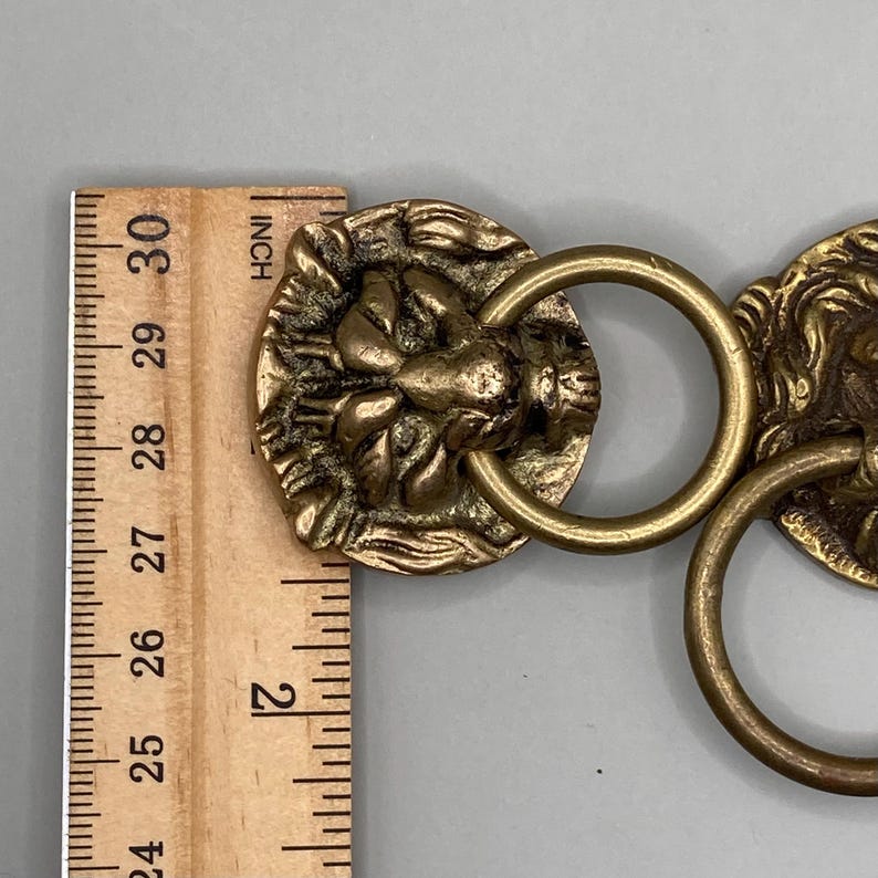Puede incluir: Dos pomos de puerta de lat&oacute;n con forma de cabeza de le&oacute;n con anillos. Los pomos miden aproximadamente 5 cm de di&aacute;metro.