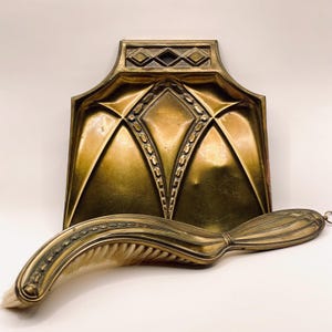 Op de afbeelding: Een koperen haardblik en een bijpassende gebogen handveger. Het haardblik is voorzien van een geometrisch ruitpatroon en een rechthoekige handgreep. De veger heeft een lange, gebogen metalen steel met een decoratieve rand en lichte haren.