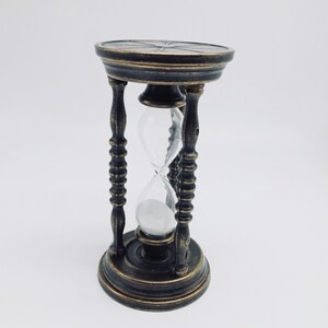 May include: A brass hourglass with a black and gold finish. The hourglass is filled with sand and has a decorative top.