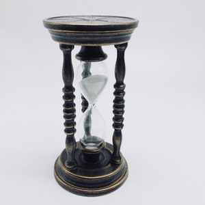 May include: A brass hourglass with a clear glass bulb and a black sand timer. The hourglass is on a white background.