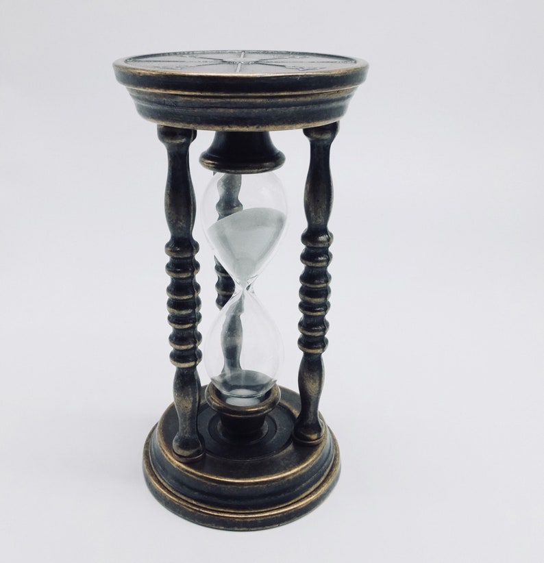 May include: A brass hourglass with a clear glass bulb and a black sand timer. The hourglass is on a white background.