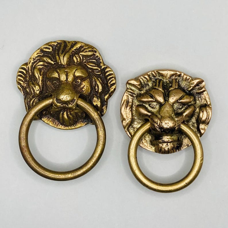 Puede incluir: Dos pomos de puerta antiguos de lat&oacute;n en forma de cabeza de le&oacute;n con tiradores de anillo. Las cabezas de le&oacute;n est&aacute;n detalladas con pelo texturizado y tienen una expresi&oacute;n feroz.