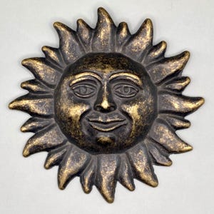 May include: A gold and black metal sun wall sculpture with a smiling face. The sun has a textured surface and a raised design.
