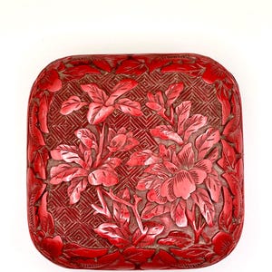 May include: A red, square-shaped box with a carved floral design. The box features a detailed pattern of flowers and leaves, with a geometric background. The edges are rounded, and the overall aesthetic is ornate and decorative.