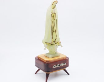 Può includere: Una statuetta bianca della Vergine Maria, in piedi su una base di legno con la scritta "FATIMA" incisa su di essa. La statuetta indossa una veste bianca e ha una corona in testa. La base ha tre gambe.