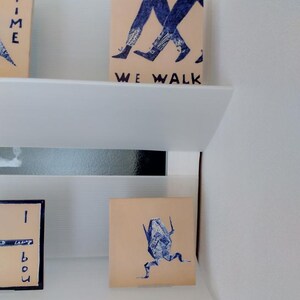 May include: A display of small, square art pieces with blue ink illustrations and text. One piece shows walking legs with the text "WE WALK". Another depicts a running figure. The art pieces are displayed on white shelves.