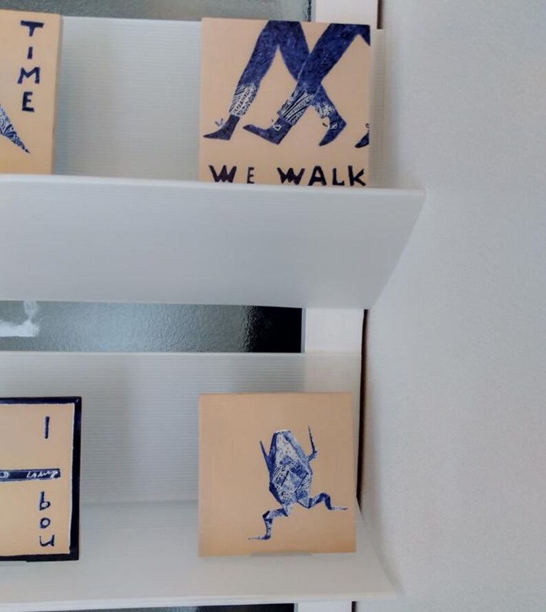 May include: A display of small, square art pieces with blue ink illustrations and text. One piece shows walking legs with the text "WE WALK". Another depicts a running figure. The art pieces are displayed on white shelves.