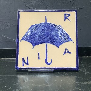 Może przedstawiać: Kwadratowa płytka ceramiczna z niebieskim wzorem parasola i literami "R", "N", "I" i "A". Parasol ma szczeg&oacute;łowy, wirujący wz&oacute;r. Płytka ma kremowe tło i niebieską obw&oacute;dkę.