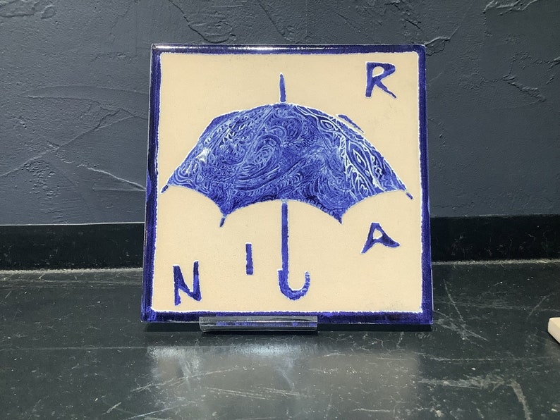 Może przedstawiać: Kwadratowa płytka ceramiczna z niebieskim wzorem parasola i literami "R", "N", "I" i "A". Parasol ma szczeg&oacute;łowy, wirujący wz&oacute;r. Płytka ma kremowe tło i niebieską obw&oacute;dkę.