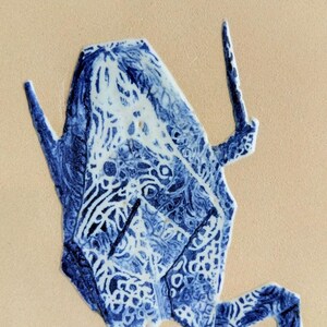 May include: A blue and white ceramic frog figurine with intricate patterns. The frog has a geometric design and is set against a beige background. The figurine's details include textured surfaces and a stylized appearance.
