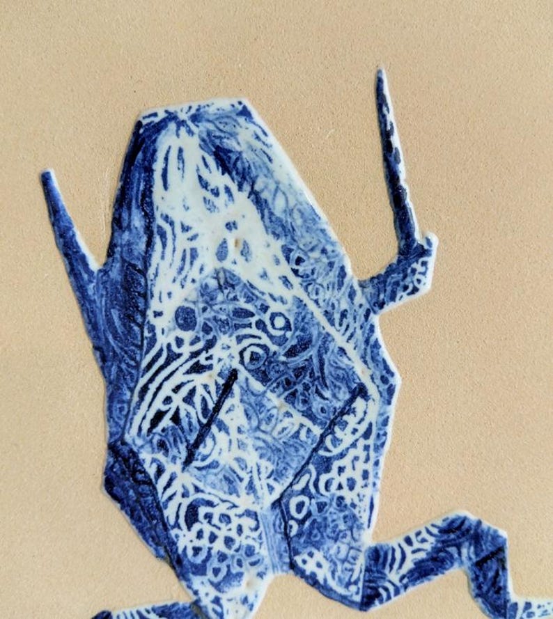 May include: A blue and white ceramic frog figurine with intricate patterns. The frog has a geometric design and is set against a beige background. The figurine's details include textured surfaces and a stylized appearance.