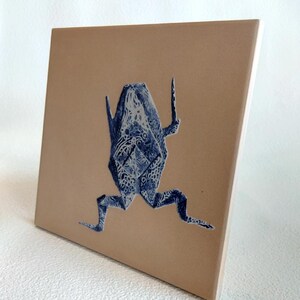 May include: A square ceramic tile featuring a blue origami frog design against a tan background. The frog is detailed with intricate patterns. The tile has a smooth, glossy finish and is suitable for home decor.