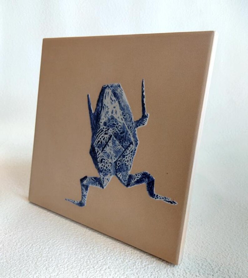 May include: A square ceramic tile featuring a blue origami frog design against a tan background. The frog is detailed with intricate patterns. The tile has a smooth, glossy finish and is suitable for home decor.