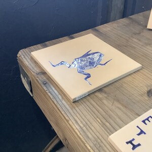 May include: Square ceramic tile with a beige background and a blue illustration of a stylized insect. The tile is displayed on a wooden surface. Additional tiles with similar designs are visible in the background.