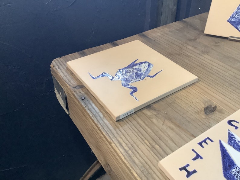 May include: Square ceramic tile with a beige background and a blue illustration of a stylized insect. The tile is displayed on a wooden surface. Additional tiles with similar designs are visible in the background.