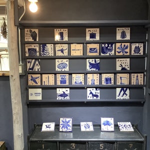 Może przedstawiać: Ekspozycja kwadratowych płytek ceramicznych z niebiesko-białymi ilustracjami i tekstem. Płytki ułożone są na p&oacute;łkach i blacie. Obrazy przedstawiają samolot, kwiat i słowo "POUND".