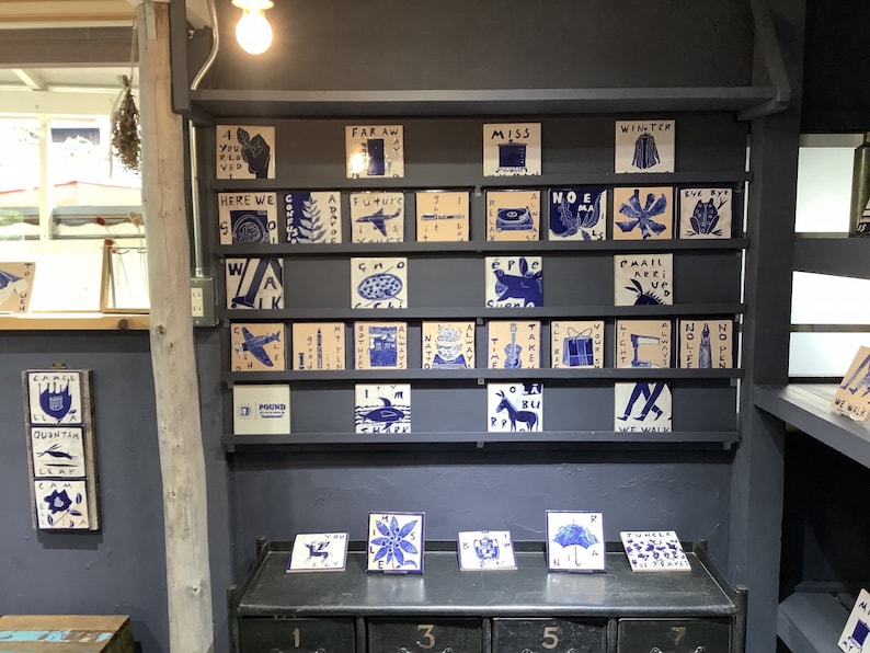 Może przedstawiać: Ekspozycja kwadratowych płytek ceramicznych z niebiesko-białymi ilustracjami i tekstem. Płytki ułożone są na p&oacute;łkach i blacie. Obrazy przedstawiają samolot, kwiat i słowo "POUND".