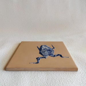 May include: A square, tan-colored coaster with a blue origami frog design. The frog is depicted in a running pose, with its legs extended. The coaster has a smooth surface and a wooden edge.