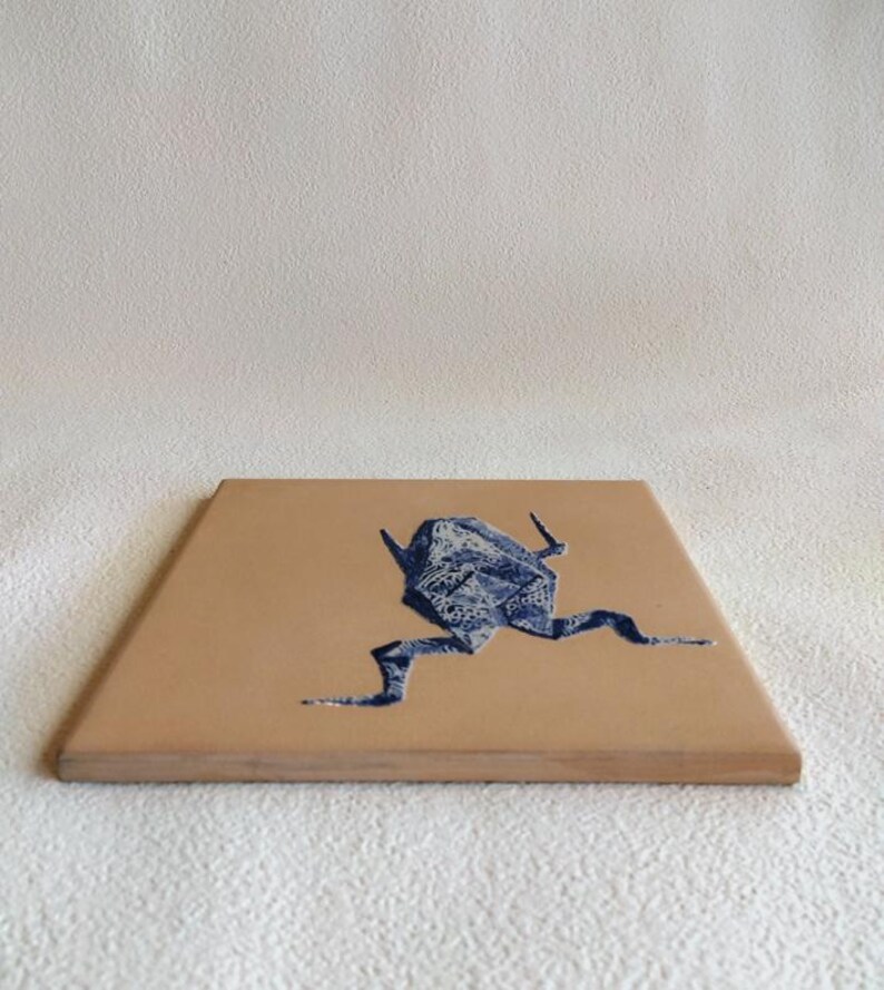 May include: A square, tan-colored coaster with a blue origami frog design. The frog is depicted in a running pose, with its legs extended. The coaster has a smooth surface and a wooden edge.