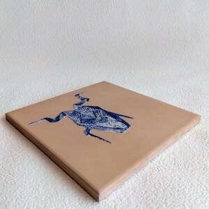 May include: A square, tan-colored tile with a blue, abstract design. The design appears to be a stylized creature or shape with intricate patterns. The tile is angled, showing its thickness and the texture of the surface.