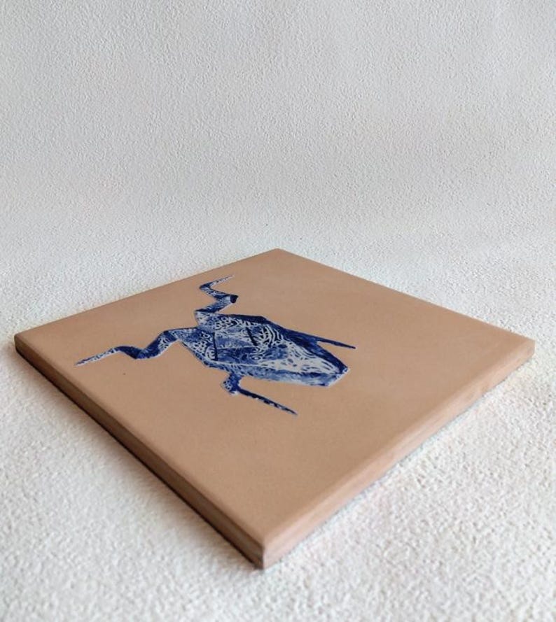 May include: A square, tan-colored tile with a blue, abstract design. The design appears to be a stylized creature or shape with intricate patterns. The tile is angled, showing its thickness and the texture of the surface.