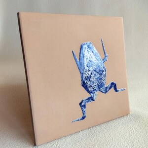 May include: Square ceramic tile with a light brown background. A blue and white abstract creature is painted on the tile. The creature has a detailed pattern and is in a running pose. The tile has a brown edge.