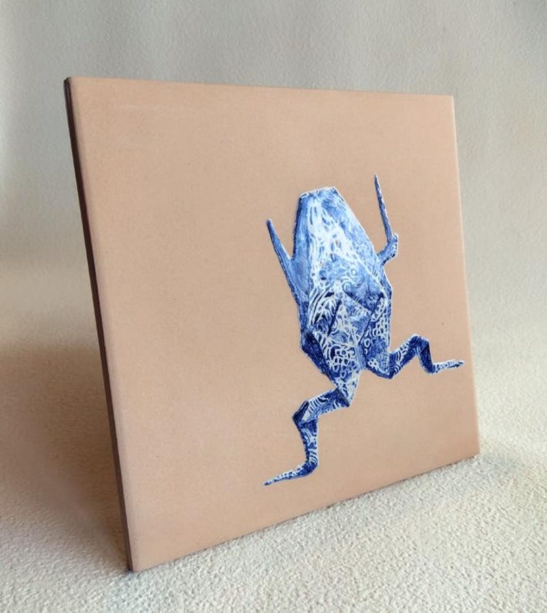 May include: Square ceramic tile with a light brown background. A blue and white abstract creature is painted on the tile. The creature has a detailed pattern and is in a running pose. The tile has a brown edge.