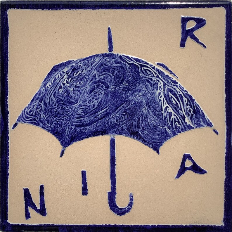 Może przedstawiać: Kwadratowa płytka ceramiczna z niebieskim wzorem parasola. Parasol ma szczeg&oacute;łowy wz&oacute;r i jest umieszczony na beżowym tle. Litery "R", "N", "I" i "A" są umieszczone wok&oacute;ł parasola. Płytka ma ciemnoniebieską obw&oacute;dkę.