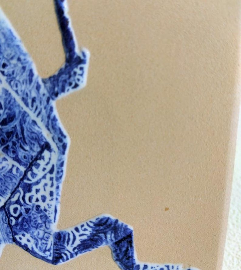 May include: Close-up of a ceramic tile with a blue and white decorative design. The design features a stylized, abstract shape with intricate patterns. The tile has a beige background.