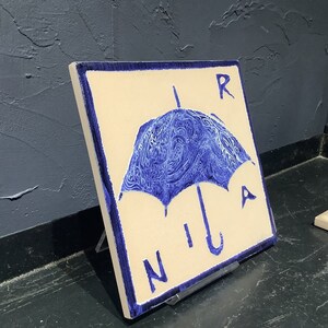 Może przedstawiać: Kwadratowa płytka ceramiczna z niebieskim wzorem parasola i napisem "RAIN" w niebieskich literach. Parasol ma szczeg&oacute;łowy, wirujący wz&oacute;r. Płytka jest w kolorze ecru z niebieską obw&oacute;dką i jest eksponowana na czarnym stojaku.