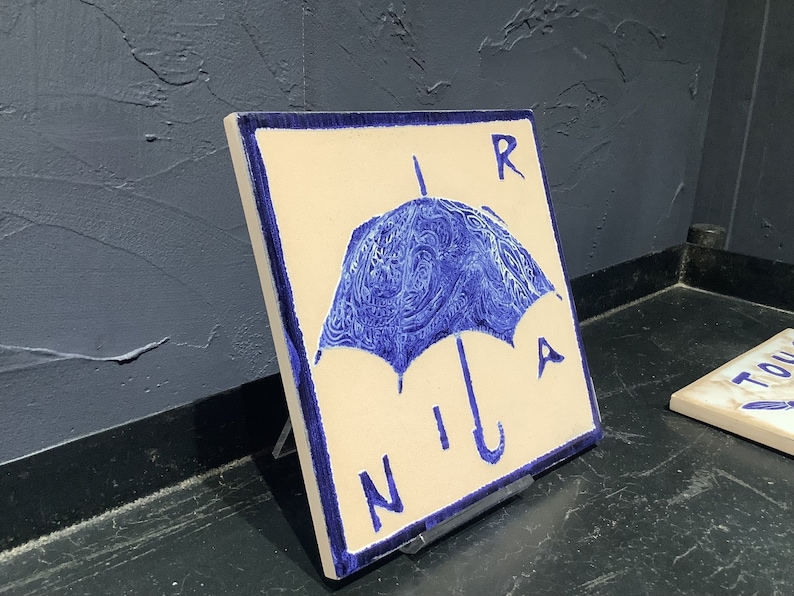 Może przedstawiać: Kwadratowa płytka ceramiczna z niebieskim wzorem parasola i napisem "RAIN" w niebieskich literach. Parasol ma szczeg&oacute;łowy, wirujący wz&oacute;r. Płytka jest w kolorze ecru z niebieską obw&oacute;dką i jest eksponowana na czarnym stojaku.
