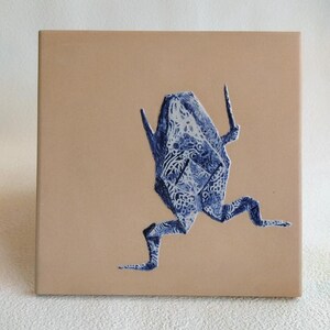 May include: A square, tan ceramic tile featuring a blue and white origami frog design. The frog is depicted mid-stride, with intricate patterns adorning its form. The artwork is centered on the tile, creating a balanced composition.