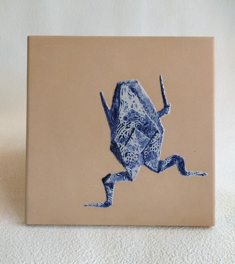 May include: A square, tan ceramic tile featuring a blue and white origami frog design. The frog is depicted mid-stride, with intricate patterns adorning its form. The artwork is centered on the tile, creating a balanced composition.
