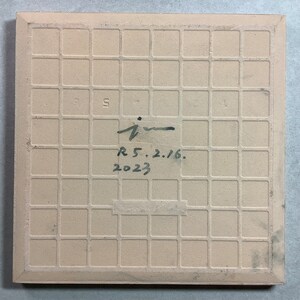 May include: A square, beige ceramic tile with a grid pattern on the back. The tile has handwritten text that reads "i", "R 5.2.16.", and "2023". The tile's edges are slightly darker.