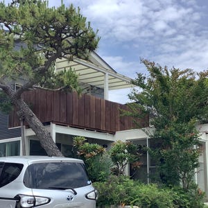 May include: A modern two-story house with a silver car parked in front. The house features a dark gray corrugated metal exterior, a wooden balcony, and a large tree growing in front. The sky is cloudy.