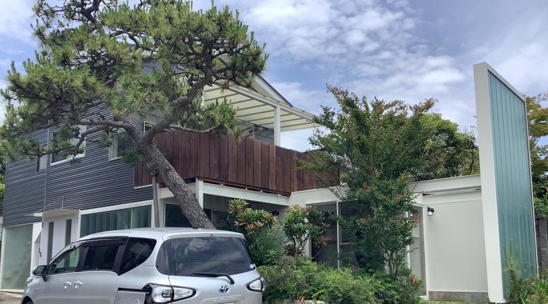May include: A modern two-story house with a silver car parked in front. The house features a dark gray corrugated metal exterior, a wooden balcony, and a large tree growing in front. The sky is cloudy.