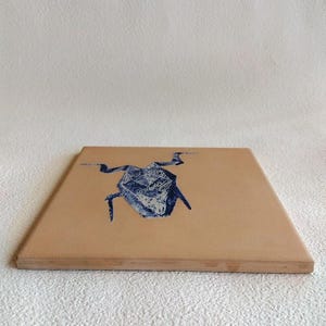 May include: A square, light brown wooden coaster with a blue and white abstract design. The design appears to be a stylized insect or creature. The coaster has a simple, clean design, suitable for various home decor styles.