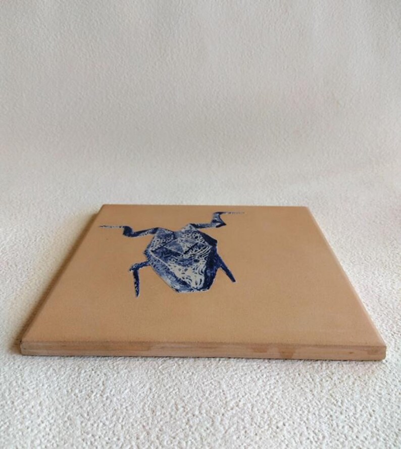 May include: A square, light brown wooden coaster with a blue and white abstract design. The design appears to be a stylized insect or creature. The coaster has a simple, clean design, suitable for various home decor styles.