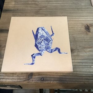 May include: Square ceramic tile featuring a blue origami frog design. The frog is detailed with a patterned, decorative print. The tile has a beige background and is displayed on a wooden surface.