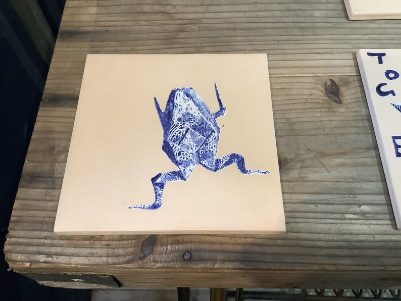 May include: Square ceramic tile featuring a blue origami frog design. The frog is detailed with a patterned, decorative print. The tile has a beige background and is displayed on a wooden surface.