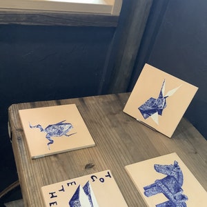 May include: Four square, beige tiles with blue illustrations of a bug, origami, a horse, and abstract shapes. The tiles are arranged on a wooden table. The text "TOGETHER" and "HER" are visible on one tile.