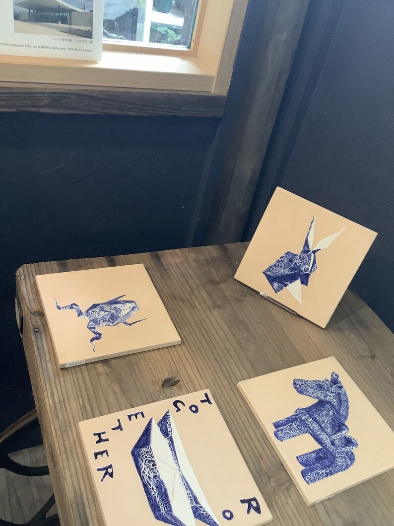 May include: Four square, beige tiles with blue illustrations of a bug, origami, a horse, and abstract shapes. The tiles are arranged on a wooden table. The text "TOGETHER" and "HER" are visible on one tile.