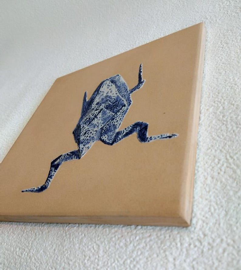 May include: A square canvas art piece featuring a blue and white origami frog design. The frog is depicted in a running pose against a tan background. The artwork has a modern, minimalist aesthetic.
